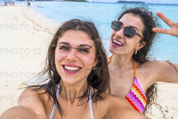 Two smiling young women taking a self portrait on a white sandy beach with turquoise ocean in the background, sharing joy and friendship during their tropical holiday