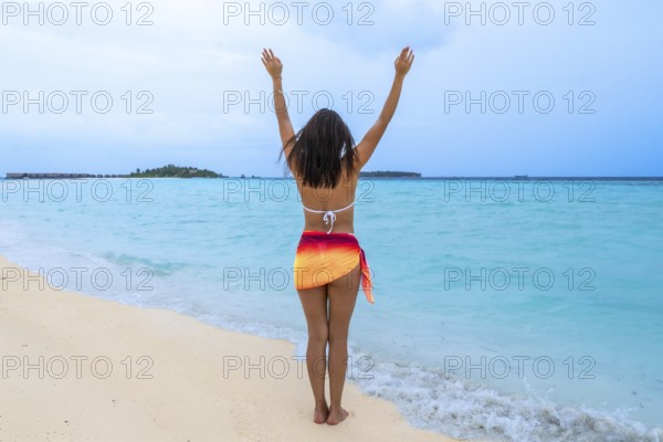 Woman standing on a white sand beach with turquoise ocean water, raising arms in joy and freedom, celebrating a luxurious tropical summer holiday in the maldives