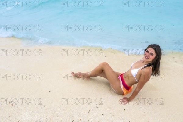 Young woman in a bikini and colorful sarong sitting on white sandy beach, smiling while relaxing by the turquoise blue ocean water during a summer holiday in the maldives