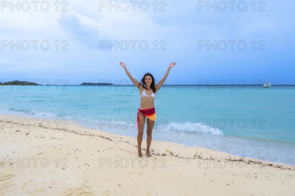 Young woman standing on a pristine white sand beach with turquoise water, raising her arms in joy and celebrating a relaxing holiday amidst the beautiful ocean scenery and distant islands