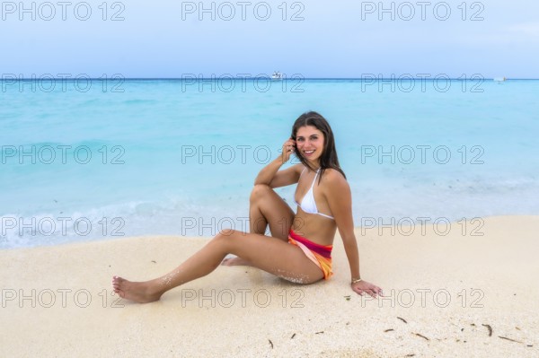 Young woman sitting on white sand beach smiling, enjoying a relaxing tropical getaway with clear turquoise ocean, distant boat and sunny blue sky on an island resort