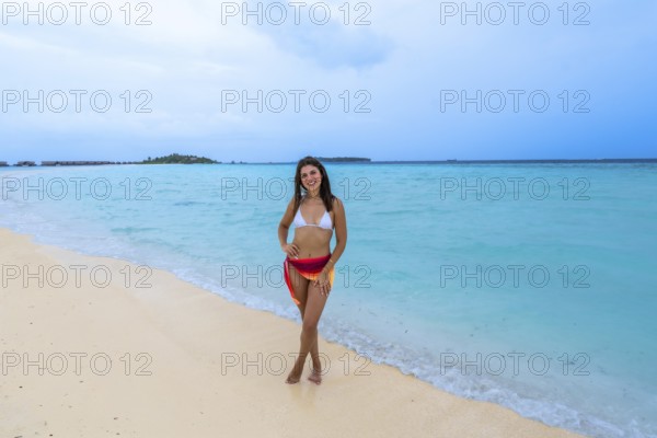 Young woman smiling while standing on a pristine sandy beach with turquoise ocean water, enjoying a tropical vacation in the maldives with overwater bungalows in the distance