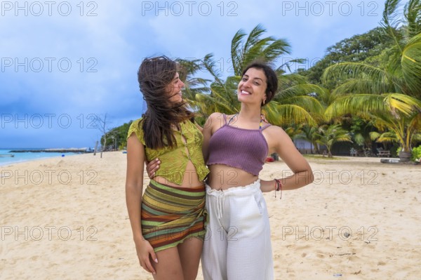 Young women friends embracing on a sunny tropical beach, smiling and enjoying a relaxing island holiday, surrounded by swaying palm trees and clear blue ocean water