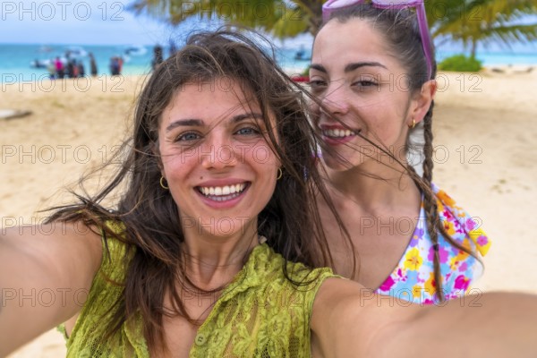 Two young happy women smiling for a close up selfie, standing on a sandy beach during a summer vacation, enjoying travel and friendship together in a tropical paradise