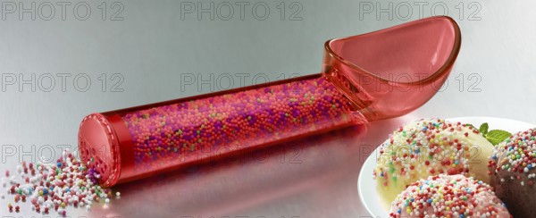 Red, transparent ice cream scoop filled with coloured sugar sprinkles, next to a plate with different types of ice cream, decorated with coloured sugar sprinkles