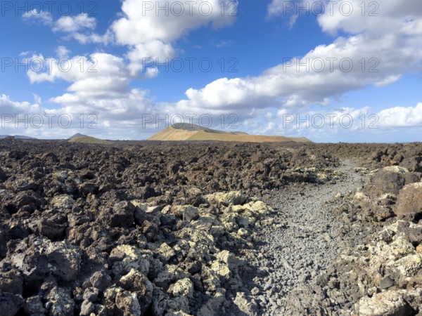 Hiking trail through lava rock to Caldera Blanca volcano against blue sky with white clouds, Mancha Blanca, Lanzarote, Canary Islands, Spain