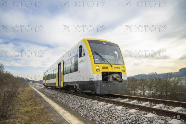 A train moves through open countryside on rails under a blue and white sky, taking off the Hermann Hessebahn, Calw, Germany