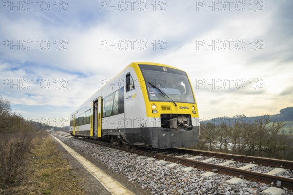 A yellow and white train travels on rails through an open landscape under a cloudy sky, taking off the Hermann Hessebahn, Calw, Germany