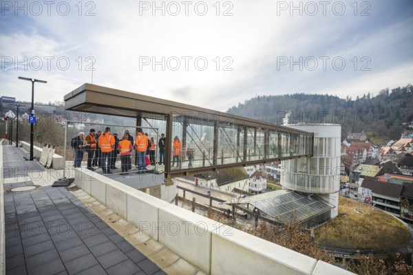 People in orange vests stand on a glass bridge with a view of a city and hills, taking off the Hermann Hessebahn, Calw, Germany