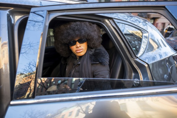 Black woman with a confident expression, wearing sunglasses and a stylish afro, is sitting in a car with the door open, reflecting urban buildings on a sunny winter day in manhattan
