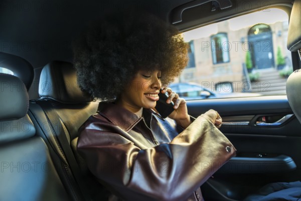 African american woman with afro hairstyle smiling while telecommunicating on a mobile phone in the back seat of a car in an urban new york city street