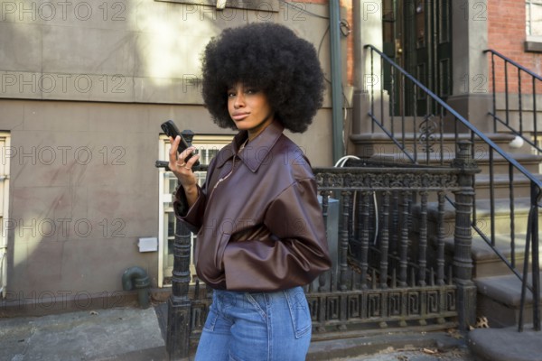Young woman with afro hair wearing a brown jacket and jeans, looking at the camera while holding a mobile phone, representing urban lifestyle and connectivity in new york city