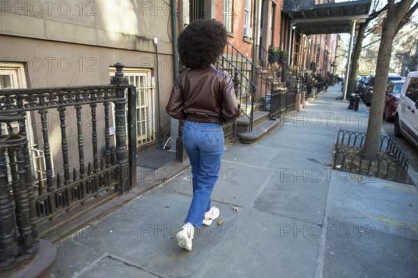 Young woman with afro hair walking along a manhattan sidewalk past brownstones, casual denim jacket and confident stride capturing urban lifestyle, autumn sunlight and architecture