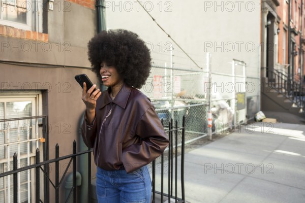 Young african american woman with afro hairstyle laughing while sending an audio message on her smartphone, enjoying urban street life on a sunny day in manhattan