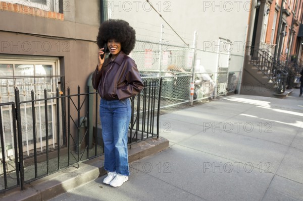 Young black woman with an afro hairstyle standing on a manhattan street, wearing a bomber jacket and jeans, laughing while having a phone conversation on her mobile device