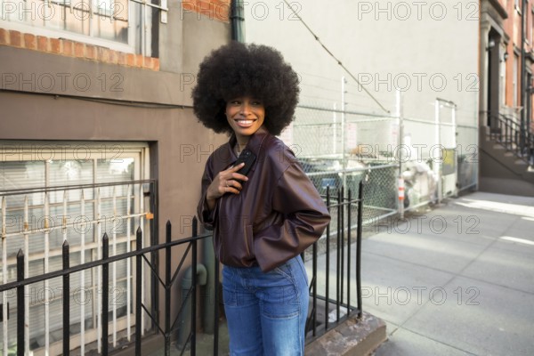 Young african american woman with a large afro hairstyle smiling, wearing a leather jacket and jeans, standing beside a fence on an urban sidewalk in new york city