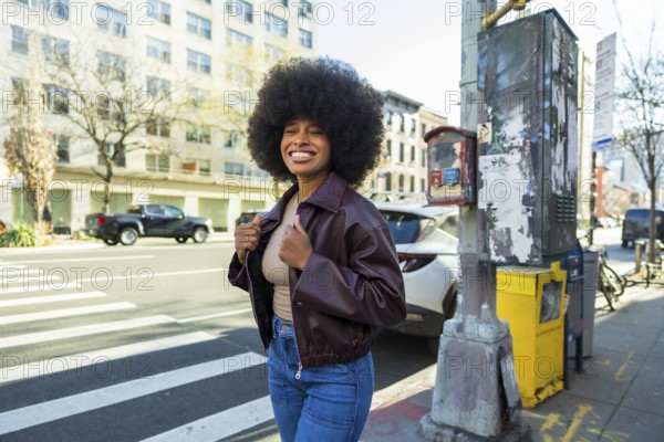 Young african american woman with a big afro hairstyle and smiling face standing on a new york city street, wearing a brown jacket and jeans, representing urban joy and diverse lifestyle