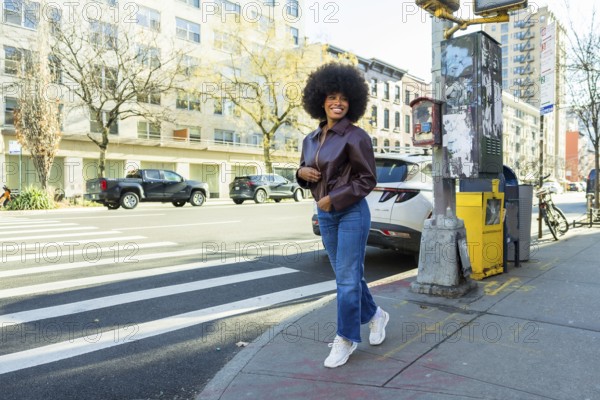 Young black woman smiling confidently, walking briskly on a manhattan city street, wearing a stylish leather jacket and jeans, capturing urban lifestyle and fashion trends