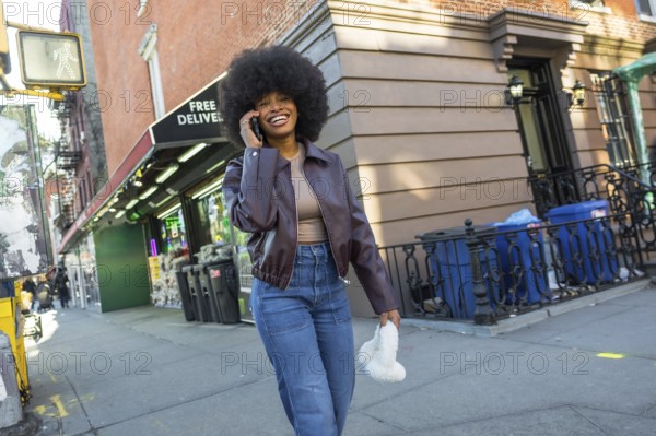 Happy young woman with an afro smiling widely, talking on her mobile phone while walking along a bustling sidewalk in manhattan, united states, enjoying urban life