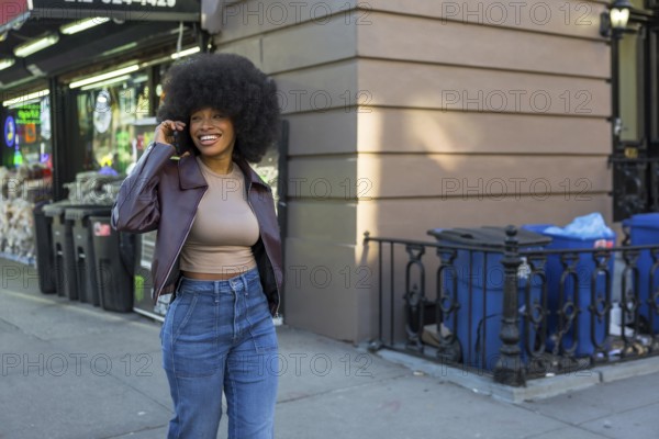 Young black woman with afro hairstyle walking manhattan sidewalk, smiling and chatting on smartphone, casual denim jacket, enjoying urban life and lively downtown energy