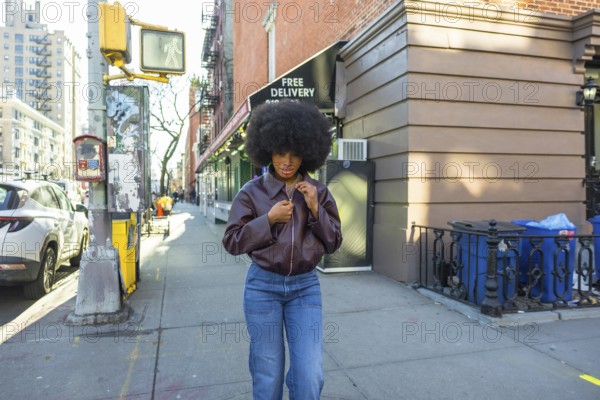 Young black woman with an afro hairstyle adjusting her brown leather jacket while standing on a manhattan street sidewalk, showcasing urban fashion in a vibrant city environment
