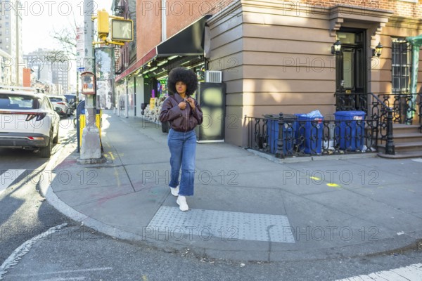 Young woman with natural afro hair walking confidently along a residential brownstone building on a sunny new york city sidewalk, showcasing urban street style in manhattan