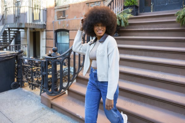 Young black woman embracing urban lifestyle while happily walking down steps of a traditional brownstone building, showcasing casual fashion in a vibrant manhattan neighborhood