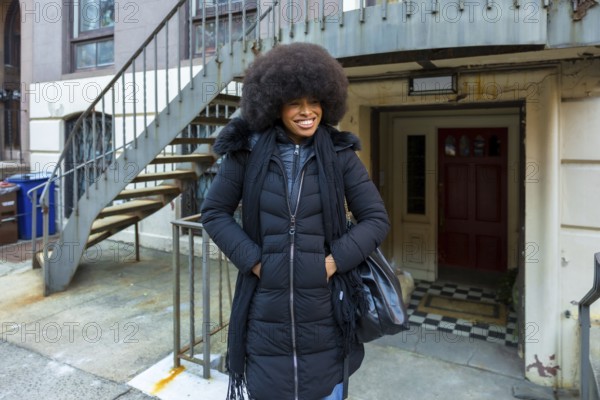 African american woman smiling while standing in front of a manhattan brownstone building. Wearing a black winter coat and scarf. Showcasing urban lifestyle and natural afro hairstyle on a city street
