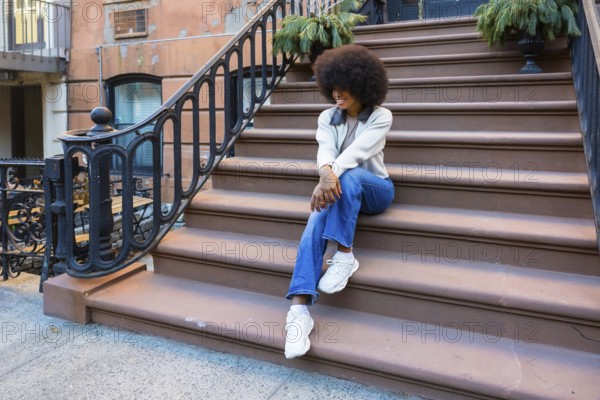 Young african american woman with an afro hairstyle sitting casually on brownstone stoop steps. Enjoying a relaxed moment in a vibrant new york city neighborhood. Embodying urban style and daily life