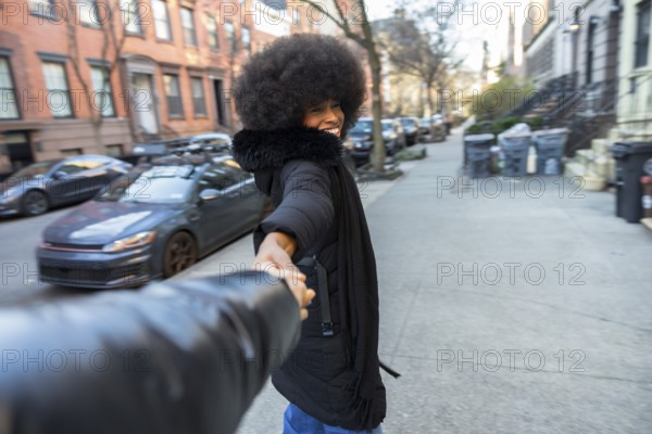 Smiling young african american woman in a winter coat leading a journey, holding hands with a partner while walking on a residential street in manhattan, embracing urban lifestyle and happiness