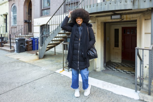 Joyful black woman with afro smiling confidently on a manhattan brownstone street in casual winter street fashionpuffer jacket, wide leg jeans and scarfvibrant urban style and confidence