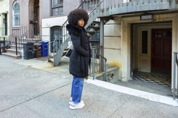 Young woman with large afro hair wearing a black winter coat and blue jeans standing on a city street sidewalk, looking attentively to the side while posing in an urban setting
