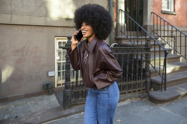 African american woman with an afro hairstyle smiling and communicating on a smartphone while standing on a sidewalk in front of a manhattan brownstone building