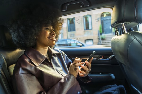 Young woman with afro hairstyle smiling, riding in the backseat of a car through new york city, using her smartphone for communication or navigation during an urban commute