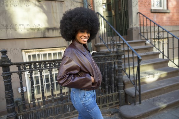Young african american woman smiling while posing with an afro hairstyle and brown bomber jacket against a classic brownstone apartment building and wrought iron railing in manhattan