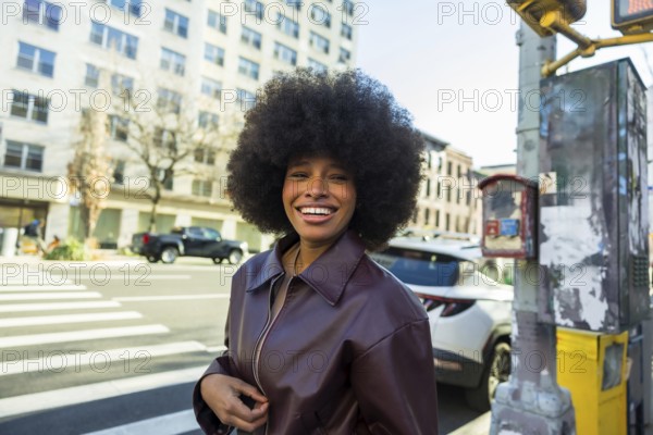 Happy young african american woman smiling brightly, standing on a new york city street with an afro hairstyle and a stylish jacket, embodying urban lifestyle and positivity