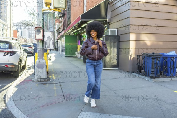 Young black woman with a vibrant afro smiling as she walks a sunny manhattan street in casual bomber jacket, wide leg jeans and sneakers, radiating confidence and urban style