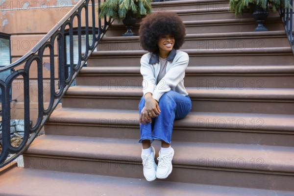 Young black woman sitting on brownstone steps in harlem, smiling and relaxed in casual street styleblue jeans, sweater and sneakers embodying urban confidence and joy