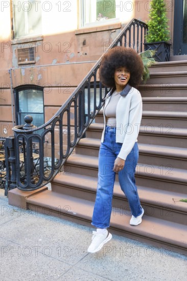 Smiling woman with afro hair descending brownstone steps in new york city, expressing joy and freedom while enjoying urban lifestyle and casual fashion