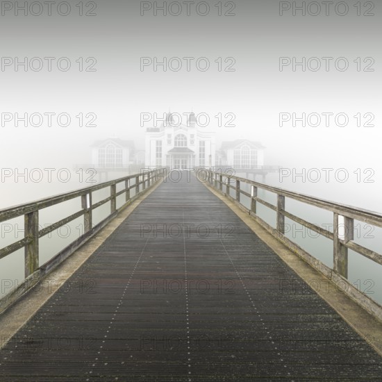 A long wooden walkway leads through thick fog to a white building, view of pier in Sellin in thick fog