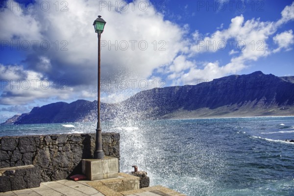 Water splashes on a stone wall with mountains and lantern, Caleta de Famara Lanzarote