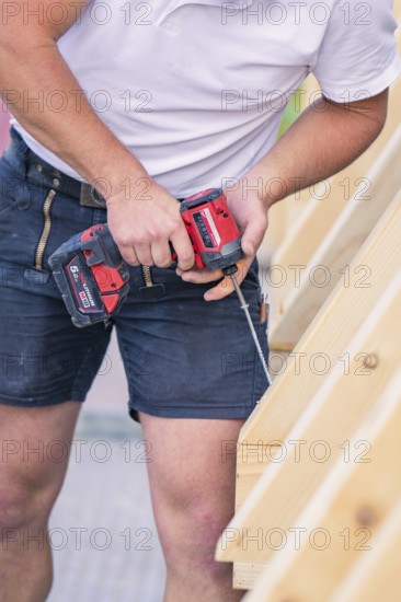 A construction worker in shorts holds a screwdriver and works on a wooden structure, carpentry, new building, Stuttgart, Germany