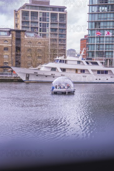 A dome boat on calm water in front of a modern marina with buildings in the background, London, England, Great Britain