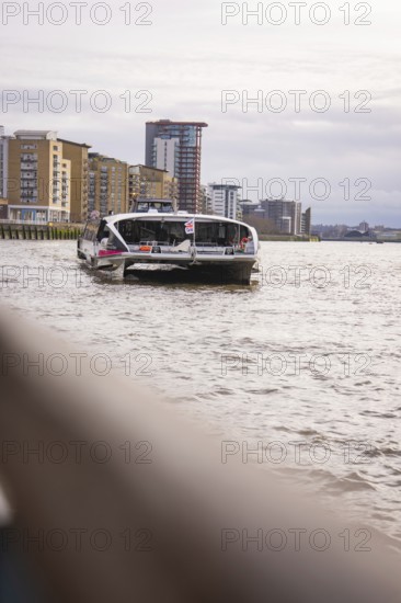 A boat on a wide river with modern buildings in the background and a cloudy sky, London, England, Great Britain