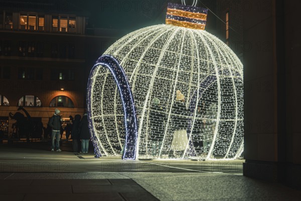 Large illuminated Christmas ball in public square with passers-by at night, London, England, Great Britain