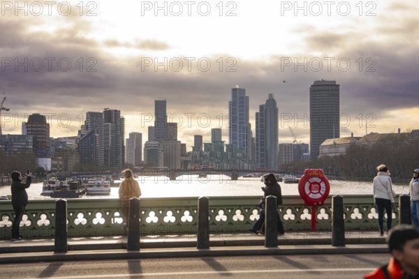 View across a bridge to the skyline of a city with river and cloudy sky, people in the foreground, London, United Kingdom