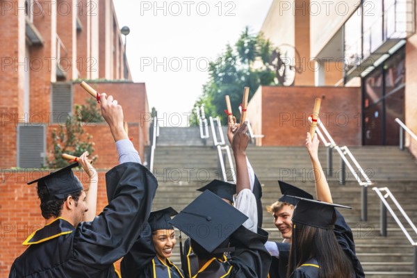 Group of diverse graduating students wearing academic caps and gowns, happily raising their diploma scrolls in celebration on a university campus after achieving their academic goal