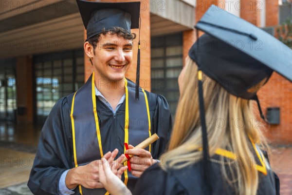 Graduating male student holding a rolled diploma, smiling and talking with a female classmate, both wearing black academic cap and gown with yellow stole on a university campus