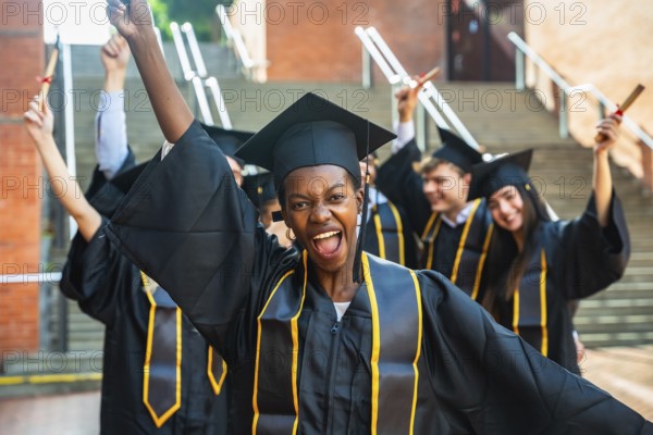 Smiling young woman posing with raised arms and holding a diploma while celebrating her academic success with diverse classmates in graduation caps and gowns