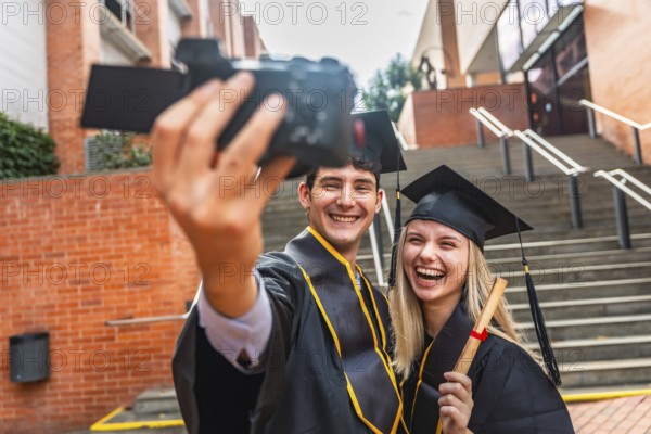 Graduating young man and woman wearing academic dress holding a diploma and filming a celebratory video on a camera, standing outdoors on campus stairs